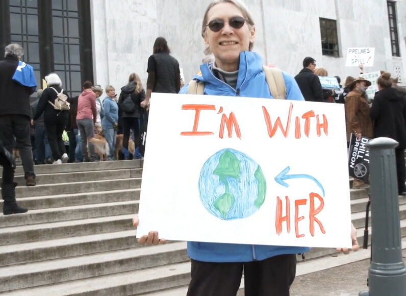 Woman stands outside with a sign that reads I'm with her and has a drawing of the earth